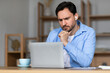 © Prostock-studio - A concentrated man is seated at a desk in a well-lit room, looking intently at his laptop screen. He wears a blue shirt and wireless earbuds, and there is a cup of coffee next to him