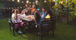 © ihorvsn - Joyful multi-ethnic young people speaking at garden party in evening. Pretty Caucasian and African American girls and guys sitting at table. Happy Asian woman. Friends raising glasses and bottles