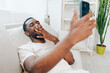 Happy African American man sitting on a black sofa, typing on his mobile phone The modern apartment is filled with a relaxing atmosphere as he enjoys some leisure time His confident smile shows his