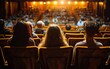 © OLGA - Audience sits in theater seats, warmly lit, watching a performance.