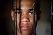 © Emvats - Portrait of a teenage prisoner staring intently into the camera behind the bars of a prison cell.
