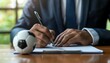 © Creative_Bringer - A person in a suit signing a document with a soccer ball on the table, suggesting a sports contract