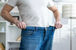 © Liubomir - Man in a white shirt showcasing significant weight loss by wearing oversized jeans in a well-lit room.