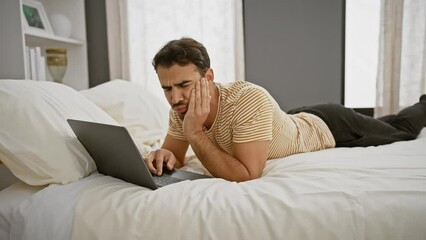 Wall Mural - A young hispanic man with a beard looks stressed while working on his laptop in a bedroom setting.