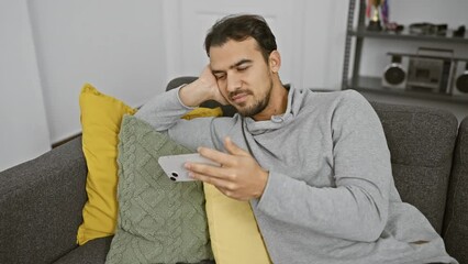 Wall Mural - A relaxed hispanic man sits on a couch at home, browsing his smartphone in a casual setting.