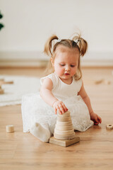 Wall Mural - Cute beautiful girl baby sitting on the floor in the children's room and playing in eco wooden toys cubes and pyramid