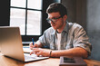 © BullRun - Concept of technology and communication, clever male copywriter in eyeglasses keyboarding on laptop computer connected to wifi, young man looking at netbook screen and writing notes for work