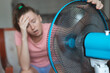 © Евгений Шемякин - Young woman using electric fan at home in living room, sitting on couch cooling off during hot weather, suffering from heat, high temperature