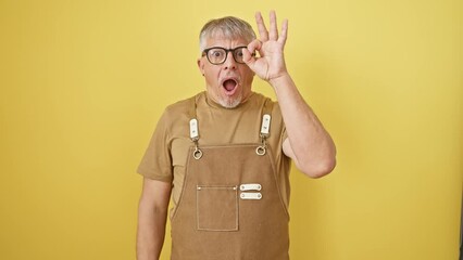 Poster - Shocked middle-aged grey-haired man in glasses and apron doing a cheeky 'ok' gesture, peeping through fingers with a surprised expression. standing against a vibrant yellow isolated background.