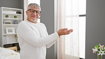 Poster - Excited middle-aged, grey-haired man cheerfully pointing aside, presenting ad in bedroom. wearing pyjamas, he is showing open palms towards copy space, happily advertising on a homey background.