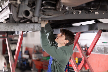 Wall Mural - Young guy mechanic in uniform repairs underbody of car in car service station