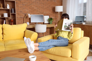 Wall Mural - Young woman reading magazine in yellow armchair at home