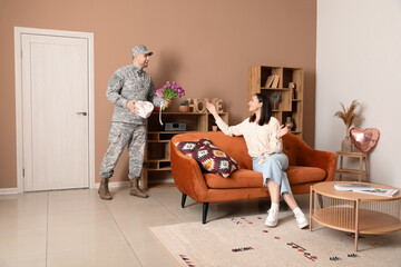 Wall Mural - Man in military uniform greeting his wife with flowers and heart-shaped gift box at home. Valentine's Day celebration