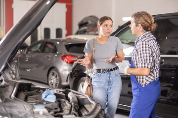 Wall Mural - Adult male mechanic advises young woman client on repairing under hood of car in car service station