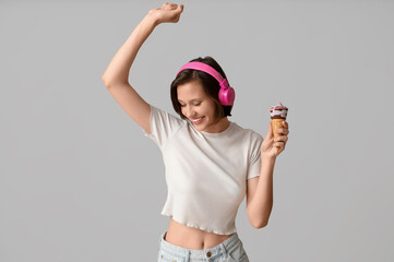 Happy young woman in headphones with sweet ice-cream in waffle cone on grey background