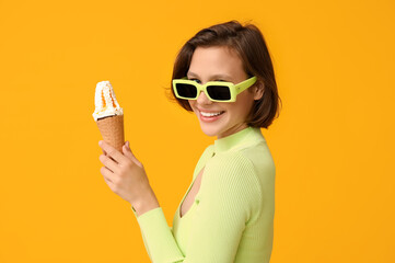 Wall Mural - Beautiful young woman in sunglasses with sweet ice-cream in waffle cone on yellow background