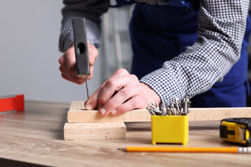 Wall Mural - Professional repairman hammering nail into board at wooden table indoors, closeup