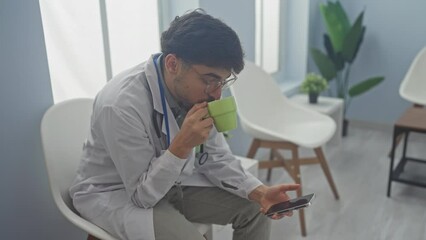 Canvas Print - A young bearded male doctor in glasses, using a smartphone and drinking coffee while taking a break in a hospital waiting room.