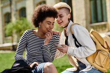 Wall Mural - Two young girls in stylish attire sitting on the grass, engrossed in phone, amidst a serene setting.