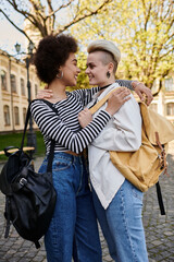 Wall Mural - Two young women, one Black and one Caucasian, embrace each other warmly while holding backpacks outdoors.