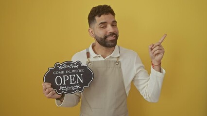 Canvas Print - Young, happy hispanic man in apron smiling and singing, pointing finger to the side over isolated yellow background