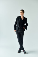 Wall Mural - A young queer man, dressed in a sleek black suit, confidently poses for the camera in a studio with a grey background.