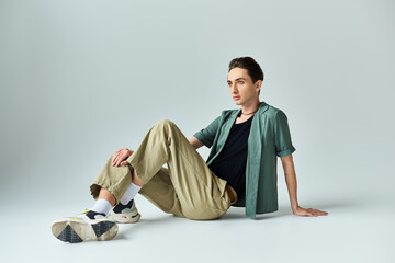 Wall Mural - A young queer person in a tan shirt and khaki pants sits on the floor in a thoughtful pose in a studio setting against a grey background.