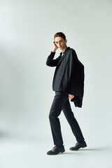 Wall Mural - A young queer person strikes a pose in a studio, donning a stylish black jacket and pants, against a grey background.