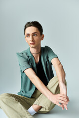 Wall Mural - A young queer person, wearing a green shirt and tan pants, sits on the floor in a reflective pose against a grey background.