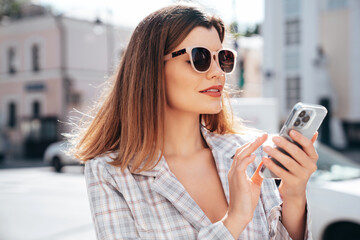 Wall Mural - Young beautiful woman in nice trendy white jacket suit. Sexy smiling model posing in street. Fashionable female outdoors. Cheerful and happy. Holds smartphone, uses phone apps, looks at screen