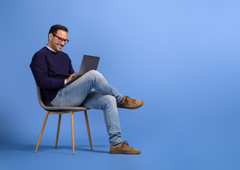 Wall Mural - confident young businessman in eyeglasses using laptop while sitting on chair over blue background