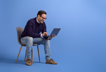 Wall Mural - smiling businessman checking online websites over laptop while sitting on chair over blue background