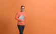 © Moon Safari - Handsome entrepreneur with hand in pocket holding laptop and smiling at camera on orange background