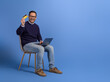 © Moon Safari - Portrait of cheerful young man with laptop and credit card sitting on chair against blue background
