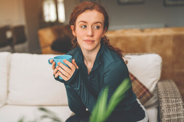 Wall Mural - cheerful redhead woman sitting on sofa, drinking coffee at home  