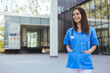 © Dragana Gordic - A joyful Caucasian nurse in blue scrubs stands confidently outside a hospital, stethoscope around her neck, with her colleagues in the background.