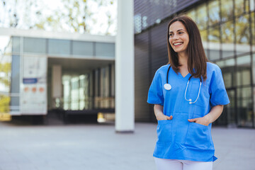 A joyful Caucasian nurse in blue scrubs stands confidently outside a hospital, stethoscope around her neck, with her colleagues in the background.