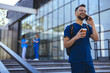© Dragana Gordic - A cheerful male healthcare worker in scrubs engages in a phone conversation outside a clinic, coffee in hand, with colleagues in the background.