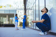 © Dragana Gordic - A weary male healthcare worker takes a moment to rest outside a hospital, his concerned colleagues engage in discussion behind him, reflecting the strenuous nature of their profession.