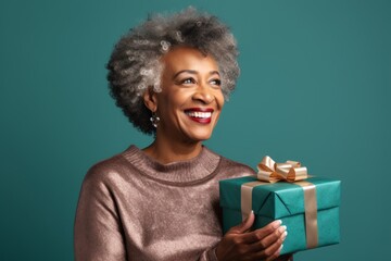 Wall Mural - Portrait of a glad afro-american woman in her 50s holding a gift over soft teal background