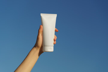 Female hand holding a white mockup tube of cream on a blue sky background. Concept of natural moisturizing, nourishing and sun protection cosmetics for skin, beauty and spa.