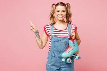 Wall Mural - Young smiling happy woman wear red t-shirt denim overalls casual clothes hold rollers point index finger aside on area isolated on plain pastel pink background. Summer sport lifestyle leisure concept.