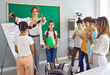 © Studio Romantic - Happy children and teacher having fun activities in classroom. Educator and students playing game in front of classroom board. Teacher holding schoolgirl by braids and classmates applauding to girl