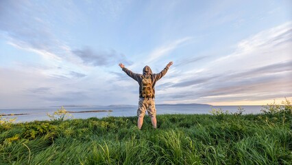 Man hiker in camo shorts and backpack on top of green hill, hands up, view on wild Atlantic way at Galway, Ireland, freedom, adventure and lifestyle concept, nature background