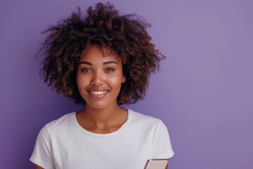 Wall Mural - Photo of happy black woman with afro hair wearing casual t-shirt holding a cellphone and smiling at camera on purple studio background