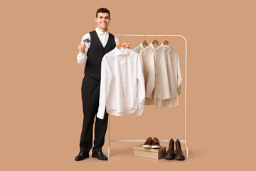 Wall Mural - Handsome young happy man holding credit card and rack with clean shirts on brown background