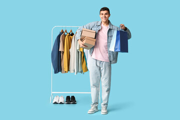 Handsome young man holding shopping bags, cardboard boxes and rack with stylish clothes on blue background