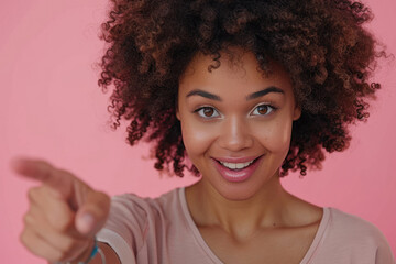 Wall Mural - Photo of a happy black woman with afro hair wearing a casual t-shirt pointing his index finger at the camera on a pink background