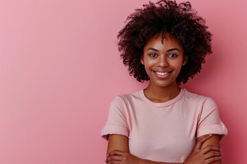 Wall Mural - Photo of a happy black woman with afro hair wearing a casual t-shirt standing with crossed hand on a pink background