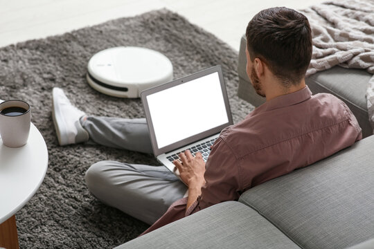 Young man with laptop and modern robot vacuum cleaner sitting on carpet at home